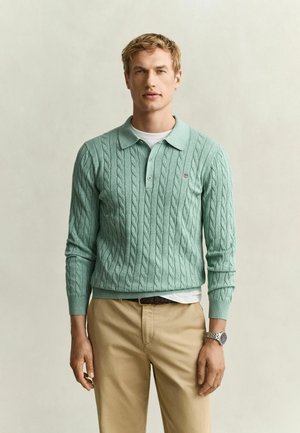 Blonder Mann trägt ein hellgrünes, grob gestricktes Langarm-Poloshirt, beigefarbene Hose und eine Armbanduhr, steht vor einem schlichten Hintergrund.
