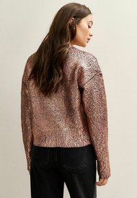 Chaqueta metálica de color rosa dorado con un acabado texturizado y agrietado. Presenta mangas largas y un corte relajado, combinada con jeans negros.