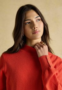 Roter Strickpullover mit hohem Kragen, mit goldenen Knopfakzenten an den Manschetten. Weiche Textur, taillierte Passform.