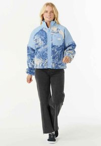 Blonde vrouw die loopt, gekleed in een blauwe fleecejas met patroon en borstzak, zwarte flared jeans en zwarte sneakers met witte zolen.