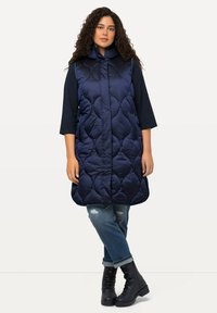 Marineblaue, gesteppte ärmellose Weste mit hohem Kragen, die eine glatte Textur, seitliche Taschen und einen abgerundeten Saum aufweist, kombiniert mit verwaschenen Jeans und schwarzen Stiefeln.