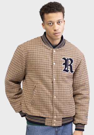 Jaket v barvi tan s houndstooth vzorom, z rebrastim mornarsko modrim ovratnikom in manšetami, zapiranje na gumbe ter velikim mornarsko modrim logotipom "R" na prsih.