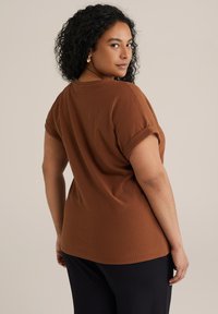 Haut brun texturé avec des manches courtes retroussées, présentant une coupe décontractée et un ourlet arrondi. Porté avec un pantalon noir, montré de profil.