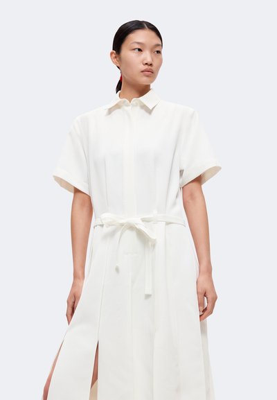 Femme aux cheveux foncés attachés, portant une robe blanche à manches courtes ceinturée avec des fentes latérales, regardant sur le côté sur un fond clair.