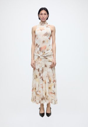 Γυναίκα όρθια, φοράει αμάνικο φόρεμα με σχέδιο λουλουδιών, ψηλό λαιμό και ένα στριφτό στοιχείο στη μέση, με μαύρα μυτερά τακούνια, ουδέτερο φόντο.