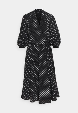 Femme portant un manteau couleur chameau par-dessus une robe à pois noirs et blancs, un sac bandoulière noir et des chaussures à talons noires, debout sur un fond uni.