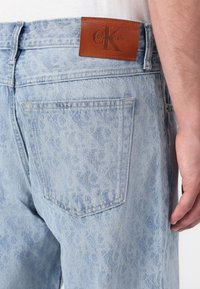 Jean en denim bleu clair avec un motif de texture subtil, une étiquette de ceinture en cuir marron avec un logo embossé, et des poches arrière standards.