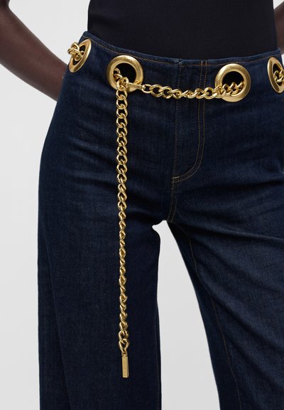 Mørkeblå jeans med store gyldne nitter ved taljen, gennemtrukket med et chunky gyldent kædebælte, der hænger ned foran på venstre side.