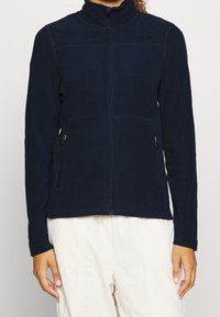 Giacca in pile blu navy con colletto alto, chiusura a zip davanti, due tasche laterali e una finitura testurizzata. Accentazione del logo discreta sulla parte superiore sinistra.