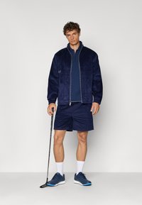 Mørkeblå kordfløyelsjakke med glidelås, matchende mørkeblå polo og shorts. Glatte sneakers med blå detaljer. Holder en golfkølle.