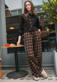 Cardigan di lana marrone con bottoni, abbinato a pantaloni a gamba larga a quadri in marrone scuro e giallo. Pantofole beige. Tavolo con tazza rossa e cornetto.