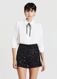 Camicia bianca con colletto impreziosito da perle e papillon nero, abbinata a pantaloncini coperti da paillettes nere lucide, con un design aderente.