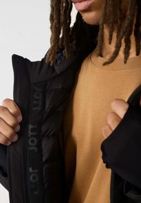 Een persoon met dreadlocks in een beige shirt trekt een zwarte gewatteerde jas open, waardoor de binnenvoering met herhaaldelijk "JOTT"-tekst zichtbaar wordt.