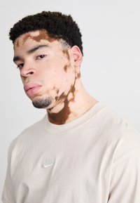 Ανοιχτό μπεζ κοντομάνικο μπλουζάκι με κέντημα του λογότυπου Nike στο στήθος. Το υποκείμενο έχει σγουρά μαλλιά και μιαDistinct Pattern Skin.