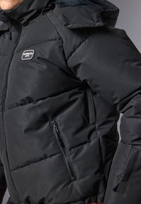 Jachetă puffer neagră cu un design matlasat, material texturat, dotată cu un buzunar cu fermoar și un patch cu logo pe piept.