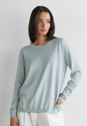 Kobieta o ciemnych włosach do ramion, ubrana w jasnoniebieski sweter z długim rękawem i białe spodnie, stoi z jedną ręką w kieszeni.