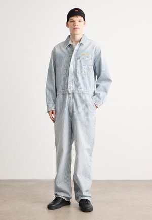 Lichtblauwe denim jumpsuit met verticale strepen, voorzien van twee voorzakken en geborduurde details, gecombineerd met zwarte sneakers.