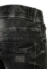 Pantalons en denim noir avec une surface texturée, des coutures visibles et un badge sur la poche arrière. Ceinture élastique en haut.