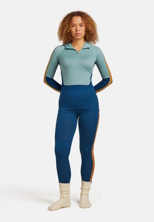 Donna con capelli ricci che indossa una maglia a maniche lunghe zip-up color teal e blu, leggings abbinati con strisce marroni e calze in maglia crema.