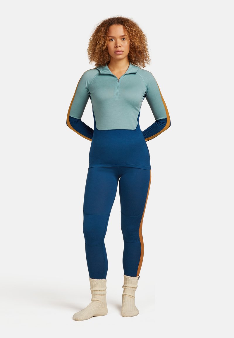 Frau mit lockigem Haar, die ein teal- und blaufarbenes Zip-up Longsleeve-Shirt trägt, passende Leggings mit braunen Streifen und cremefarbene Strumpfsocken.