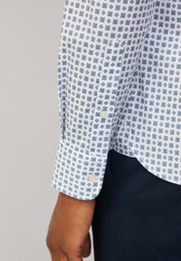 Biała koszula z niebieskim geometrycznym wzorem, z mankietami na guziki i gładką teksturą. Posiada zaokrąglony dół i widoczne guziki na rękawie.