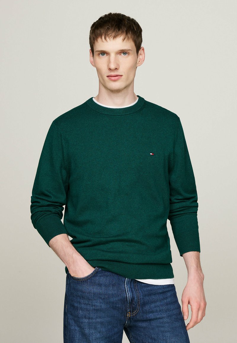 Maglione verde a maglia con colletto rotondo, polsini a coste e un piccolo logo decorativo. Abbinato a jeans blu, indossato in modo informale con le mani nelle tasche.