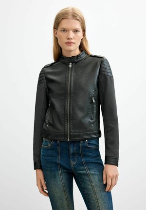 Giovane donna dai lunghi capelli biondi che indossa una giacca di pelle nera con zip e jeans blu, in piedi davanti a uno sfondo semplice.