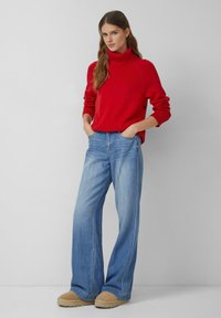 Czerwony sweter z golfem, luźne niebieskie dżinsy i beżowe wsuwane buty. Sweter ma prążkowane mankiety; dżinsy charakteryzują się prostym krojem i wyblakłą teksturą.