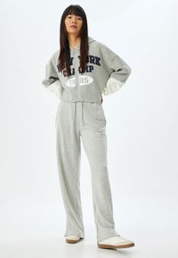 Cappuccio grigio corto con scritte blu, presenta maniche oversize. Abbinato a pantaloni della tuta grigi coordinati. Sneakers bianche con dettagli beige.