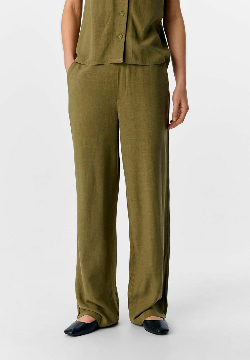 Pantalons larges vert olive en tissu léger et texturé avec une taille élastique. Associé à un haut à boutons assorti et des chaussures plates.
