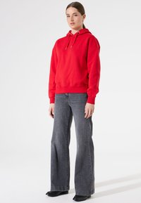 Sudadera roja con capucha y cordones, combinada con jeans acampanados grises y zapatos negros. Textura suave y corte relajado, con un detalle mínimo.