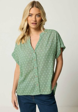 Mujer con cabello rubio largo que lleva una camisa verde de manga corta con botones y vaqueros azul oscuro con una mano en el bolsillo.