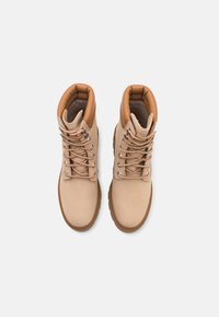 Botas de tobillo de ante beige con un cuello acolchado marrón claro, cordones planos, ojales plateados y una suela de goma marrón texturizada. Vista superior.