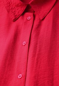 Chemise rouge vif au toucher doux, avec un col évasé et une patte de boutonnage centrée. Les boutons sont ronds et rapprochés.