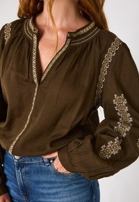 Blusa marrone a maniche lunghe con ricami decorativi lungo il collo e le maniche. Tessuto morbido, vestibilità comoda e chiusura a bottoni sul collo.