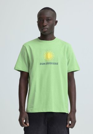 Homme à la peau foncée portant un t-shirt vert clair avec un motif de soleil jaune et le texte « SUN SEEKERS », debout devant un fond clair uni.