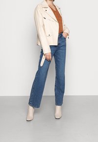 Cremefarbene Leder-Motorradjacke mit Reißverschlusstaschen, kombiniert mit einem braun gemusterten Oberteil und blauen hoch taillierten Jeans. Beigefarbene Knöchelstiefel runden den Look ab.