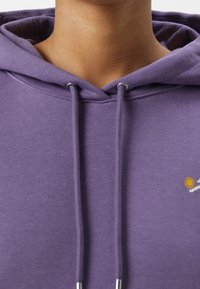 Fioletowa bluza z kapturem z troczkiem, srebrnymi elementami i małym żółtym haftem kwiatu z przodu. Miękka tekstura, swobodny design.