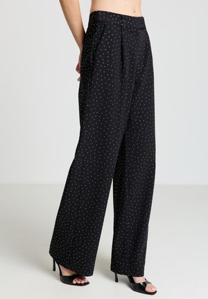Pantalon large noir à pois blancs, doté d'une taille ajustée et d'un devant plissé. Le tissu présente une texture lisse et un éclat subtil.