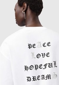 Witte sweatshirt met gedurfde zwarte en zilveren tekst op de achterkant: "VREDE," "LIEFDE," "HOOPVOL," en "DROMEN," met verschillende lettertypen.