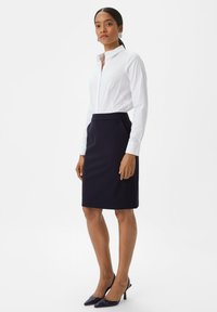 Witte blouse met kraag en marineblauwe kokerrok, met een slanke uitstraling. De rok heeft een gladde textuur en is knielang, gecombineerd met donkere hakken.
