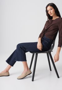 Frau mit langen dunklen Haaren sitzt seitlich auf einem schwarzen Stuhl und trägt eine dunkelbraune, geknöpfte Bluse, dunkelblaue, gekürzte Jeans und beige Schlupfschuhe.