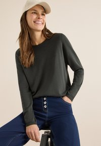 Ciemnozielona bluzka z długim rękawem i okrągłym dekoltem, do tego niebieskie dżinsy z wysokim stanem z trzema dużymi guzikami z przodu.