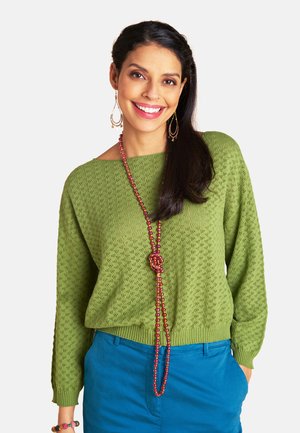 Frau lächelt, trägt einen grünen Strickpullover, eine lange rosa Perlenkette, große Ohrringe, blaue Hose und ein buntes Armband, Hand in der Tasche.