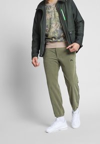 Dark green jacket with a zip pocket, camo-patterned sweatshirt, olive sweatpants with logo, and white sneakers. Soft textures and relaxed fit.