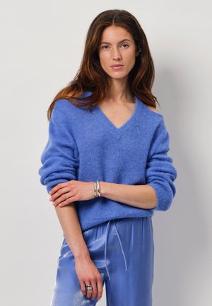 Blauwe V-hals trui met omgeslagen mouwen, gemaakt van een zachte textuurstof. Gecombineerd met lichtblauwe satijnen broek. Zilveren armband en ring accenten.