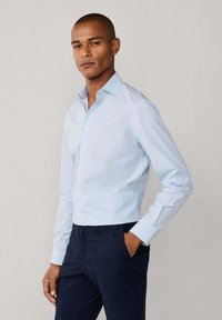 Svjetloplavi košulja dugih rukava s dizajnom na dugme, mekani materijal i šiljati ovratnik. Uparen s tamnoplavim hlačama.