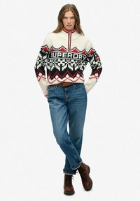 Pull en motif noir et blanc avec des accents rouges, présentant un col montant et un logo, associé à un jean bleu et des bottes en cuir marron à lacets.