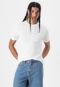 Tricou alb din bumbac, cu guler rotund și mâneci scurte. Are un mic logo brodat pe piept. Este asortat cu blugi din denim albastru deschis.