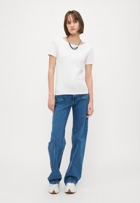 Hvit kortermet topp kombinert med blå flagrende jeans. Tilbehøret inkluderer en sølvkjedehalskjede. Innstillingen er enkel og minimal.